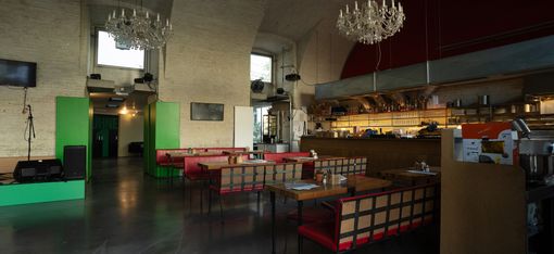 Large dining room with several tables and chairs, chandeliers, and open kitchenette in the background