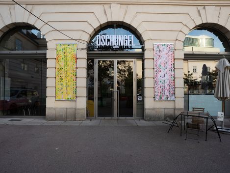 Helle Fassade mit hohen Glasfenstern. In der Mitte der Fassade befindet sich eine Tür über der der Schriftzug DSCHUNGEL WIEN angebracht ist.