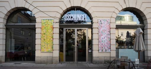 Bright facade with tall glass windows. In the middle of the facade is a door above which the word DSCHUNGEL WIEN is written.
