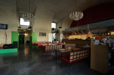Large dining room with several tables and chairs, chandeliers, and open kitchenette in the background