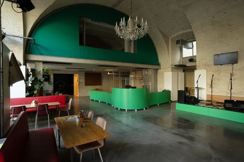 Large room with green bar, chandelier, red benches, wooden table, and stage with microphone stands and screen.