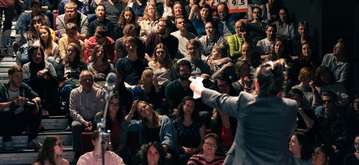 Das Foto zeigt Menschen, die im Publikum sitzen. Sie schauen auf eine Frau, die auf der Bühne steht. Neben ihr steht ein Mikrofon.