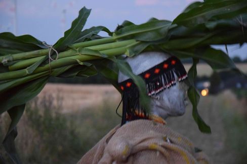 Figure with white head and black headpiece with red dots carrying large green leaves on shoulders outdoors at dusk