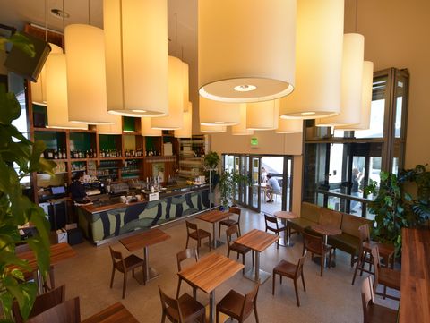 Innenraum eines Cafés mit Holztischen, Stühlen, einer Bar mit Regalen und mehreren runden Deckenleuchten