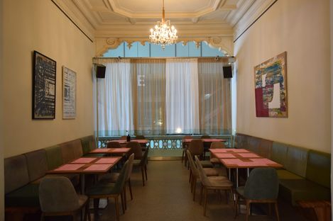 Narrow café with two rows of tables and chairs, long benches along the walls, a chandelier on the ceiling, and curtains in front of a window with a balcony