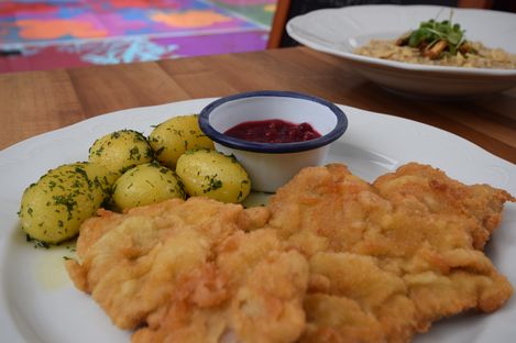 White plate with breaded schnitzel, small parsley potatoes, and a bowl of lingonberry sauce on wooden surface