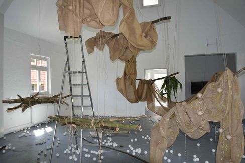Room with white walls and windows containing hanging and lying installations made of wood, fabric, and ropes including a ladder and many small white balls on the floor