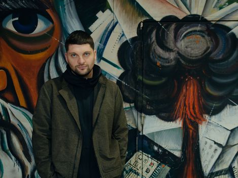 Man wearing a hooded jacket stands in front of a wall with colorful graffiti depicting a face and an explosion.