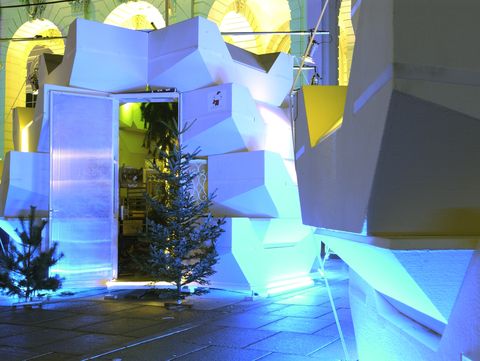 Detailansicht einer begehbaren, geometrischen Installation mit blauer und gelber Beleuchtung; kleine Tannenbäume stehen vor dem Eingang.