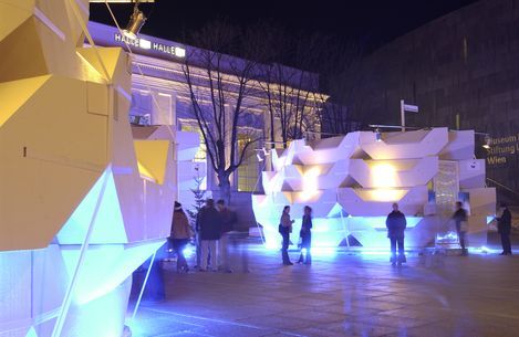 Nighttime view of MuseumsQuartier with illuminated geometric installations; visitors standing and walking between light objects in front of historic architecture.