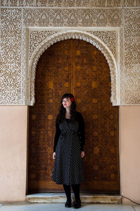 Frau in gepunktetem Kleid steht vor einer kunstvoll verzierten Holztür mit ornamentaler Architektur.