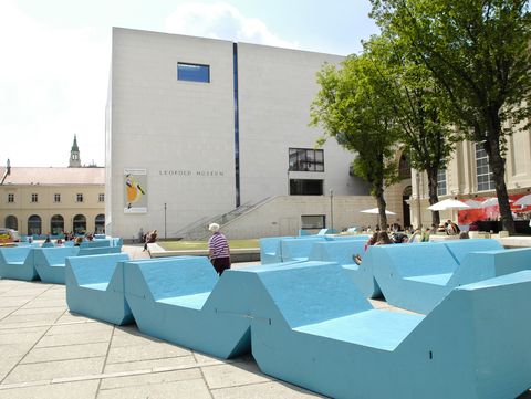 Türkise MQ-Sitzmöbel im Innenhof des MuseumsQuartier Wien vor dem Leopold Museum, mit Besucher:innen und schattenspendenden Bäumen an einem sonnigen Tag.
