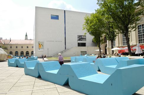 Turquoise MQ seating furniture in the courtyard of MuseumsQuartier Vienna in front of the Leopold Museum, with visitors and leafy trees on a sunny day.