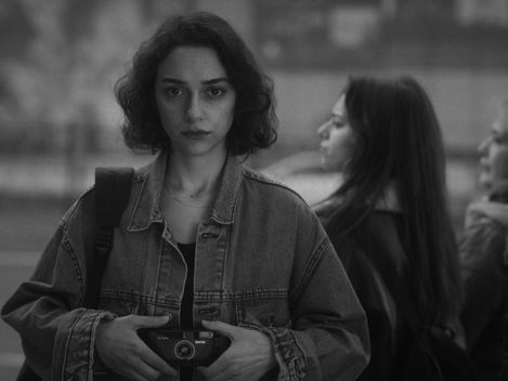 Young woman with dark shoulder-length hair wearing a denim jacket holding a camera at chest level looking directly at the camera with two blurred women in profile in the background.