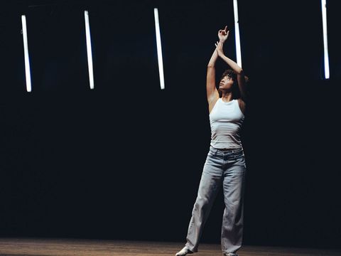 Woman in white tank top and jeans standing with raised arms against dark background with vertical light bars