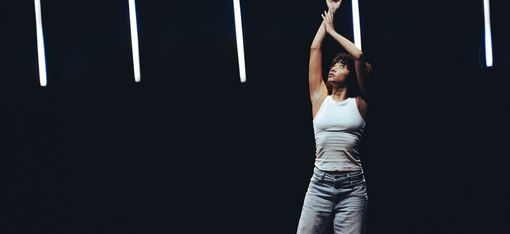 Woman in white tank top and jeans standing with raised arms against dark background with vertical light bars