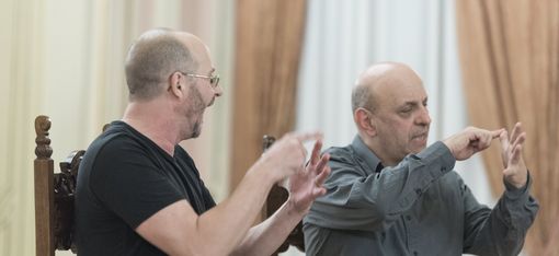 The artists are sitting side by side on chairs and gesturing.
