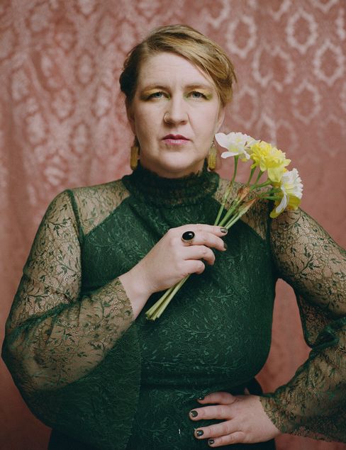 Person with updo hairstyle wearing a long-sleeved lace top holding a small bouquet of flowers against a patterned background