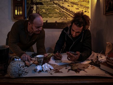 Zwei Personen sitzen an einem Tisch in einem atmosphärisch beleuchteten Raum und arbeiten konzentriert an Zeichnungen und Notizen, umgeben von Requisiten und historischen Objekten.