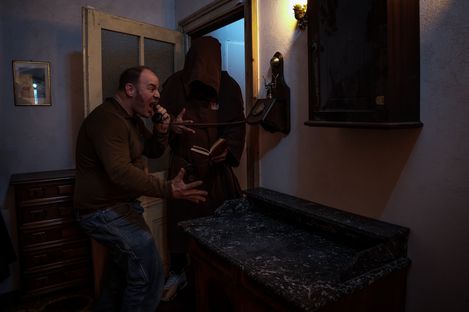 A person speaks animatedly into a wall-mounted telephone while another person in a dark hooded robe stands nearby reading from a book, in a theatrically lit interior space.
