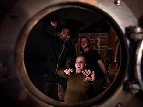 Three people pose smiling behind a circular metal opening in a dark, staged room.