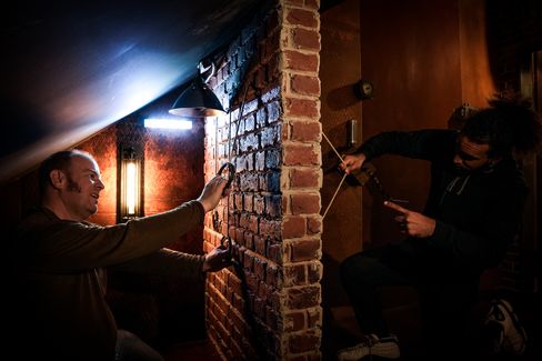 Two people work together on a brick wall with ropes and mechanical elements in a theatrical interior setting.