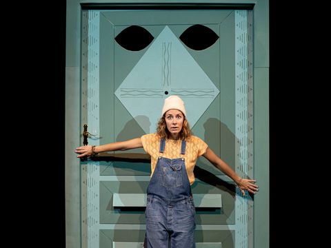 A child stands with a questioning and astonished facial expression in front of a blue door. The door has eyes, a nose, and the mail slot serves as a mouth.