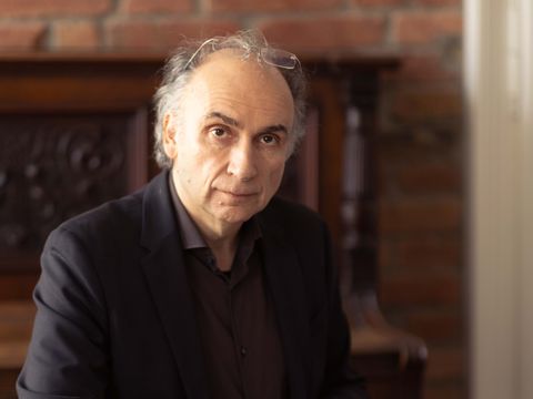 Älterer Mann mit grauem Haar sitzt an einem Holztisch vor einem Klavier und hält einen Stift über einem aufgeschlagenen Buch