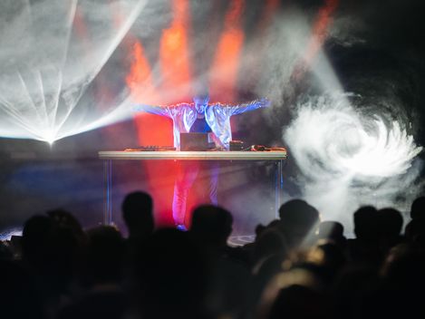 Person mit ausgestreckten Armen steht hinter einem Tisch mit elektronischem Equipment auf einer Bühne mit Nebel und bunten Lichteffekten vor Publikum.