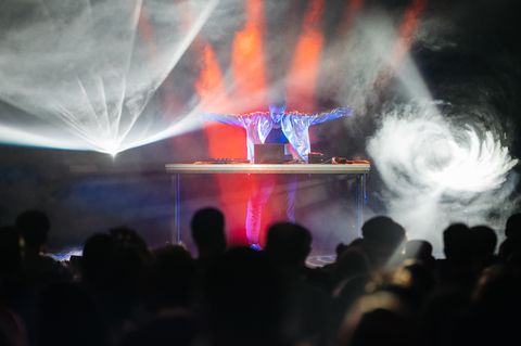 Person mit ausgestreckten Armen steht hinter einem Tisch mit elektronischem Equipment auf einer Bühne mit Nebel und bunten Lichteffekten vor Publikum.