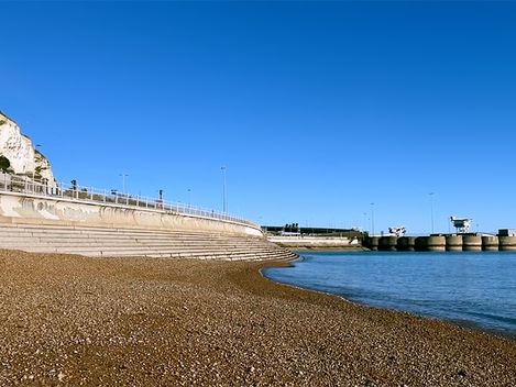 In der oberen Hälfte sieht man den blauen Himmel, auf der rechten Seite ist ruhiges blaues Meer, ein Steg und eine Strandpromenade bis zur linken Seite, auf der linken Seite sind weiße Klippen und in der Mitte Kieselstrand zu sehen.