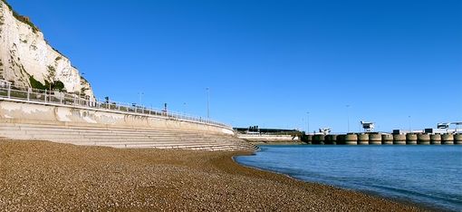 In der oberen Hälfte sieht man den blauen Himmel, auf der rechten Seite ist ruhiges blaues Meer, ein Steg und eine Strandpromenade bis zur linken Seite, auf der linken Seite sind weiße Klippen und in der Mitte Kieselstrand zu sehen.