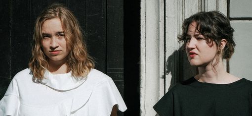 Zwei Frauen stehen nebeneinander vor einer Wand, die linke trägt ein weißes Oberteil mit Rüschen, die rechte ein schwarzes Oberteil mit kurzen Ärmeln.