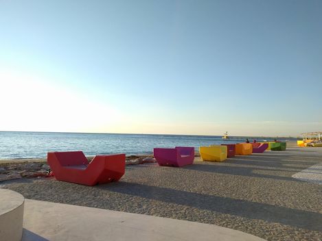 Bunte Enzi-Sitzmöbel stehen entlang einer Promenade direkt am Meer, mit Blick auf Wasser und Horizont.
