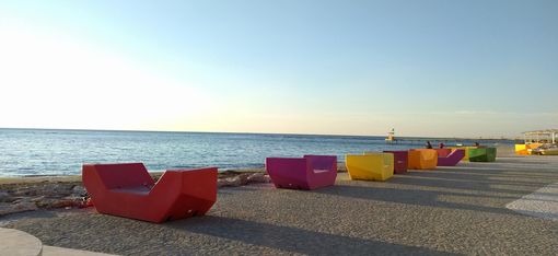 Bunte Enzi-Sitzmöbel stehen entlang einer Promenade direkt am Meer, mit Blick auf Wasser und Horizont.