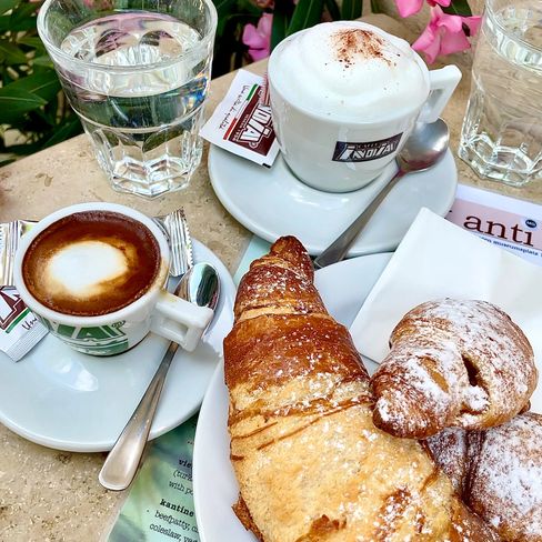 Two croissants on a plate next to a cup of cappuccino, an espresso cup, and two glasses of water on a café table.