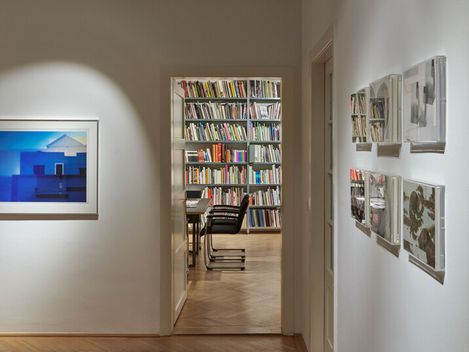 Blick durch einen Flur mit Parkettboden in einen Raum mit Bücherregal und Stühlen, an der linken Wand ein beleuchtetes Bild, an der rechten Wand mehrere gerahmte Fotos.