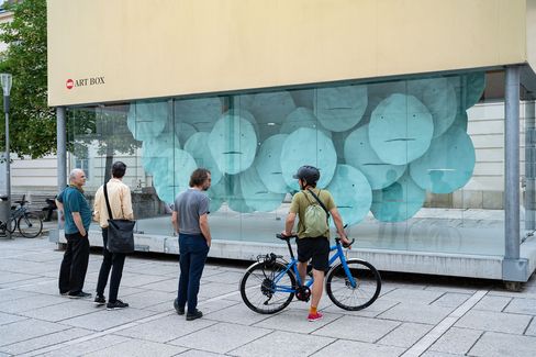Four people stand in front of a glass wall with an installation of large round blue discs, one person with a bicycle wearing a helmet and backpack