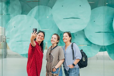 Three visitors take a selfie in front of the installation “EMOTION” in the MQ Art Box with turquoise floating face-like forms.