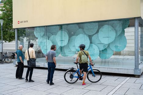 Four people stand in front of a glass wall with an installation of large round blue discs, one person with a bicycle wearing a helmet and backpack