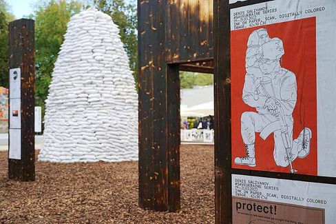 Sculpture made of white sandbags next to wooden frames with posters, one showing a drawn kneeling person holding a child and a rifle against a red background.