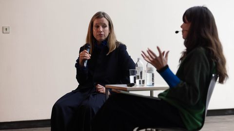 Das Bild zeigt zwei Frauen, die auf Stühlen in einem Raum sitzen und sich unterhalten. Eine Frau trägt eine schwarze Jacke und scheint zu sprechen...