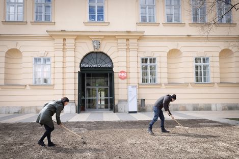 Two people with rakes work on a bare patch of ground in front of the MuseumsQuartier entrance.