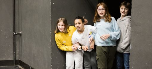 In der Nische eines Raumes vor einer offenen Tür stehen 4 Teenager, die gespannt auf einen Teil des Raumes im Vordergrund blicken, als würden sie auf ihren Auftritt warten.