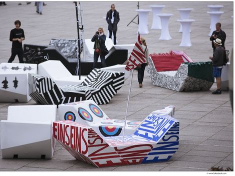 Outside urban scene with geometric, colorful benches called MQ Enzis, each featuring bold designs and patterns. People stand around, adding a lively, artistic vibe.