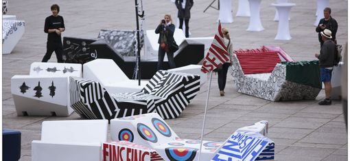 Outside urban scene with geometric, colorful benches called MQ Enzis, each featuring bold designs and patterns. People stand around, adding a lively, artistic vibe.