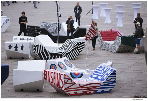 Outside urban scene with geometric, colorful benches called MQ Enzis, each featuring bold designs and patterns. People stand around, adding a lively, artistic vibe.