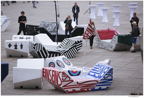 Outside urban scene with geometric, colorful benches called MQ Enzis, each featuring bold designs and patterns. People stand around, adding a lively, artistic vibe.