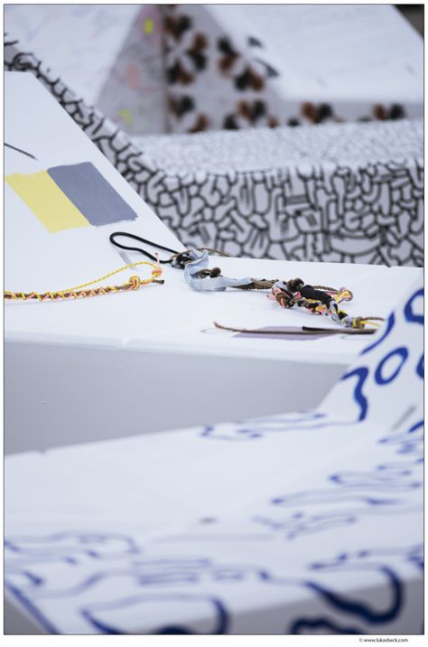 A person sits painting black, abstract circular patterns on a white, angled structure. The background is a bright, yellow-and-white striped tent.