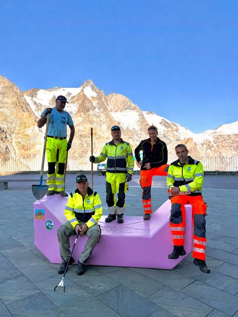Five staff members in workwear pose on and beside a pink Enzi against an Alpine mountain backdrop.